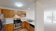This single-floor residence offers a practical and comfortable layout designed for everyday living. The kitchen is equipped with warm honey-toned wood cabinetry, generous counter space, a stainless steel electric range with an overhead vent hood, a double-basin sink, and a dishwasher. Recessed and flush-mount lighting keeps the space well-lit, while light hardwood-style flooring adds a clean finish underfoot.

The living areas are finished with neutral-toned carpet throughout, complemented by warm wood baseboards and trim that provide a cohesive look across the space. A structural column adds architectural character to the main living area, which benefits from generous natural light filtering in through large windows. One room features a wide window overlooking a tree-lined outdoor scene, allowing sunlight to fill the space throughout the day.

A hallway connects the main living areas to the private spaces, including a bedroom and bathroom. The bathroom is outfitted with a full tub-and-shower combination, a wood-trimmed vanity with a countertop sink, tile flooring, and chrome hardware and fixtures. The overall palette throughout the residence is light and neutral, creating a bright, open ambiance. The thoughtful layout ensures easy flow between the kitchen, living, and sleeping areas.