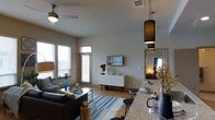 This single-story space in Euless, Texas, showcases a thoughtfully designed open-concept layout that connects the living, dining, and kitchen areas into one cohesive environment. The living area features light wood-style flooring, a contemporary sofa, a striped area rug in blue and neutral tones, and a ceiling fan that adds both function and style. Large windows fitted with blinds invite natural light throughout the space, creating a bright and welcoming atmosphere. A white mid-century modern media console serves as a focal point along the main wall, topped with a wall-mounted flat-screen TV and decorative accents. The kitchen area transitions seamlessly into the living space, anchored by a granite or stone-finished counter with bar seating. Sleek black pendant lights hang above the counter, adding contrast and visual interest. A mirror positioned near the hallway enhances the sense of depth and openness. The neutral wall palette and consistent flooring throughout contribute to a clean, unified aesthetic, while carefully placed décor elements such as florals and tableware add warmth and personality to the overall design.