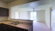 This single-floor residence showcases a thoughtfully designed open-concept layout that seamlessly connects the kitchen and living areas. The kitchen features granite-style countertops with a double-basin sink, dark cabinetry, and a built-in dishwasher. A wide pass-through opening between the kitchen and living area enhances the sense of openness and allows for easy interaction between spaces.

The living area is bright and inviting, benefiting from generous natural light that streams in through a large sliding glass door or window leading to a private balcony. A ceiling fan with a light fixture is centrally positioned in the living room, providing both comfort and functionality. The carpeted flooring in the living area adds warmth and comfort underfoot, while the neutral wall tones create a clean, versatile backdrop suitable for a variety of interior styles.

The textured ceiling treatment adds subtle visual interest throughout the space. The balcony beyond the living area offers an outdoor extension of the living space, with views of the surrounding environment. Overall, the layout is efficient and well-lit, making excellent use of the available space while maintaining a comfortable and functional living environment.