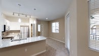 This single-story residence at 935 Northstar in San Marcos offers a thoughtfully designed open-concept layout that seamlessly connects the kitchen and living areas. The kitchen is equipped with stainless steel appliances, including a full-size refrigerator, and features a peninsula with a light-toned marble-style countertop that provides additional prep space and a natural divide between the two areas. Track lighting and a pendant light above the peninsula add functional illumination, while white cabinetry contributes to a clean, contemporary aesthetic.

The living area extends naturally from the kitchen, finished with wall-to-wall carpeting in a neutral tone that adds warmth and comfort underfoot. Smooth, neutral-toned walls and recessed lighting keep the space feeling bright and cohesive. Multiple windows, fitted with horizontal blinds, allow natural light to filter in while maintaining privacy. A glass-paneled door provides additional light and access to the exterior.

A mirrored door visible in the background hints at additional storage or closet space, and a hallway leads to the remaining areas of the unit. The overall ambiance is clean, modern, and move-in ready.