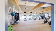 Located at 7100 E Mississippi Ave in Denver, CO, this fitness center offers a thoughtfully designed single-floor space that combines functionality with an inviting aesthetic. The facility is defined by its dramatic vaulted ceilings adorned with prominent exposed natural wood beams, which lend a warm, architectural character to the room and create a sense of openness and height throughout.

The main workout area is spacious and open, featuring light wood-tone flooring that contributes to the bright, airy atmosphere. Large floor-to-ceiling windows and glass doors along one wall flood the space with abundant natural light while providing views of the outdoor surroundings — ideal for an energizing workout environment. Multiple flat-screen televisions are mounted throughout for entertainment during exercise.

The facility is well-stocked with a wide variety of cardio equipment, including treadmills, ellipticals, and stair climbers, as well as spinning bikes for cycling workouts. A full-length mirror wall on one side visually expands the space and supports strength and form-focused training. Additional amenities include a cable machine, medicine balls, stability balls, and a dedicated free-standing punching bag area.

A secondary alcove area provides additional room for stretching or functional fitness, complete with built-in cubby storage and a freestanding boxing dummy. The overall ambiance is modern, clean, and motivating — an ideal fitness amenity for the Denver community.