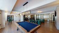 This well-appointed amenity lounge is designed to offer a dynamic and social atmosphere across a single, open-concept floor. The space is anchored by a full-size pool table positioned beneath a striking geometric pendant light fixture with Edison-style bulbs, creating an immediate focal point upon entry. Adjacent to the game area, a comfortable lounge zone features tufted emerald green sofas and accent chairs arranged around a rustic wood coffee table, all centered on a textured stone fireplace wall with a built-in linear gas fireplace and a large wall-mounted television above. Rich hardwood-style flooring flows seamlessly throughout, lending warmth and continuity to the space. A dedicated wet bar area stands out with a bold navy blue ceiling, marble-veined countertops, glossy black tile backsplash, and pendant globe lighting — paired with velvet bar seating for a sophisticated, intimate feel. A dining and meeting area with bar-height tables and upholstered green chairs provides a functional space for gatherings. Floor-to-ceiling glass-partitioned rooms line one side, offering private spaces with natural light filtering in from large windows. High windows along the perimeter bring in ample daylight, while layered ambient and accent lighting ensures a welcoming atmosphere at any hour.