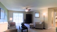 This single-floor space centers around a carpeted living room designed for comfort and functionality. The room is illuminated by natural light streaming through a large sliding glass door fitted with vertical blinds, which opens to an outdoor patio where outdoor seating is visible. A ceiling fan with an integrated light fixture is centrally mounted on the textured ceiling, providing both air circulation and supplementary lighting. A tall floor lamp in the corner adds warm ambient light to the space. The living area accommodates a sofa, a patterned armchair with a matching ottoman, and a dark wood coffee table, creating a well-arranged seating layout. A tall, dark wood armoire serves as a prominent storage or media piece along the back wall, complemented by a decorative gold-framed mirror mounted nearby. Neutral-toned walls and beige carpeting contribute to a calm, cohesive aesthetic throughout. A decorative vase with foliage and a bowl centerpiece on the coffee table add subtle character to the room's overall presentation.