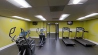 This fitness center offers a well-equipped workout space designed for convenient, everyday exercise. The room features bold yellow and white accent walls that create an energetic atmosphere, complemented by a dark rubberized floor ideal for gym use. Recessed panel lighting embedded in the drop-tile ceiling provides bright, even illumination throughout the space. The cardio equipment lineup includes treadmills, an elliptical trainer, and a stair-climbing machine, all arranged to allow comfortable spacing between users. Two wall-mounted flat-screen televisions — one positioned on each side of the room — offer entertainment during workouts. A mirrored glass door at the far end of the space adds depth to the room while providing access to an adjoining area. The overall design is utilitarian and functional, prioritizing ease of use and efficient use of space. The single-floor layout ensures straightforward accessibility, making it a practical amenity for regular fitness routines.