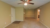 This single-floor residence offers a straightforward and functional layout designed for comfortable everyday living. The main living area is open and spacious, featuring neutral beige walls, plush wall-to-wall carpeting, and a classic five-blade ceiling fan with a built-in light fixture that provides both air circulation and ambient lighting. A textured ceiling adds a traditional architectural detail throughout the space. The entryway is clearly defined by a transition from wood-look flooring to carpet, and features a solid white front door with deadbolt hardware, providing a secure and welcoming entry point. A decorative chair rail runs along the perimeter of the living area, adding a subtle design element to the walls. Recessed lighting in the background provides additional illumination deeper into the space. The open sightlines connect the living area to the kitchen, which is partially visible and features cabinetry, allowing for a connected feel between the main living and cooking spaces. A hallway visible on the far right suggests access to additional rooms beyond, completing this practical and livable single-floor layout.