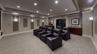 This dedicated home theater and screening room offers a refined cinematic experience within a single, thoughtfully designed space. The room is anchored by multiple rows of plush, dark leather recliner chairs arranged in a stadium-style layout, each equipped with built-in cup holders for added convenience. The focal point of the space is a large flat-panel display set within a deep navy-blue accent wall, flanked by in-wall speakers and framed by crisp crown molding that adds an elegant architectural touch.

The walls are finished in warm neutral tones and feature strategically placed acoustic panels, ensuring optimal sound quality throughout the room. Recessed ceiling lighting, complemented by soft wall sconces, creates a warm and immersive ambiance that can be tailored to any viewing occasion.

Along one side of the room, a handsome built-in credenza with rich dark wood cabinetry and a granite countertop provides ample surface space for décor and serving. A beverage refrigerator is integrated nearby, making the space fully self-contained for entertaining. Plush, patterned carpet covers the floor, enhancing both comfort and acoustics. Double glass-paned entry doors add a polished finishing touch, reinforcing the sense of a purpose-built, premium screening environment.
