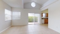 This single-floor residence is defined by its dramatic vaulted ceilings in the main living area, which rise to an impressive height and are punctuated by a distinctive circular skylight that floods the space with natural light. Large-format light tile flooring extends throughout the living and kitchen areas, contributing to the open, airy feel of the interior.

The spacious living area features multiple windows fitted with vertical blinds and a sliding glass door that opens onto a balcony overlooking lush greenery, providing pleasant outdoor views and additional natural light. Architectural interest is added through arched and stepped ceiling details that frame transitions between spaces.

The kitchen is well-appointed with warm honey-toned wood cabinetry arranged in a U-shaped layout, dark laminate countertops, a stainless steel sink, and white appliances including a range, dishwasher, and refrigerator. A window with vertical blinds provides natural light to the workspace.

Additional rooms branch off from the main living area, with carpeted bedrooms offering a softer feel. A generously sized walk-in closet with wire shelving systems provides substantial storage. The entry foyer is clearly defined, and the overall layout promotes a functional, connected flow throughout the home.