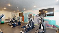 Located at 251 W 12th St in Ogden, Utah, this fitness center offers a comprehensive exercise environment within a single, open floor plan. The space is thoughtfully laid out to accommodate a variety of workout routines, with durable dark rubber flooring throughout providing both safety and functionality.

The room is well-stocked with cardio equipment, including multiple treadmills, elliptical machines, stationary bikes, and a stair climber. A standout feature is the eye-catching full-wall cityscape mural, which adds a vibrant, urban aesthetic to the interior and is complemented by a wall-mounted flat-screen television. A second television is also positioned for visibility across the equipment area.

Recessed lighting in the textured ceiling ensures the space is well-lit, while horizontal windows along one wall invite natural light and outdoor views. The utility area includes a dual-station water fountain with a bottle-filling option, promoting hydration convenience. Additional amenities include medicine balls and a stability ball for supplementary training.

The space also features a secondary door offering access to an adjacent area, as well as a utility room housing mainwater shutoff and electrical systems. Overall, the fitness center is designed to be practical, energetic, and fully functional for daily exercise needs.