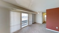 This single-floor space features an open living area designed to maximize both comfort and natural light. A wall of floor-to-ceiling sliding glass doors fitted with vertical blinds serves as a standout architectural element, flooding the interior with daylight while providing access to an outdoor balcony with urban views beyond. The tray ceiling above adds a subtle sense of dimension and architectural interest to the main living area. Neutral-toned wall-to-wall carpeting extends throughout, contributing to a warm and cohesive aesthetic. A ceiling fan is centrally positioned, ensuring year-round comfort and air circulation. The layout flows openly from the main living space toward an adjoining hallway, which leads to additional areas of the unit. Soft, warm lighting accents the connecting corridor, reinforcing an inviting ambiance. The overall design is clean and understated, with a neutral color palette of whites and beiges that provides a versatile backdrop suited to a variety of furnishing styles. The combination of generous natural light, open layout, and practical features makes this a functional and comfortable living environment.