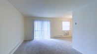 This single-floor space features an open living area finished with neutral-toned walls and plush carpeting underfoot, creating a clean and understated aesthetic. Natural light is a prominent feature, with a large sliding glass door dressed in vertical blinds serving as a focal point along one wall, allowing generous daylight to fill the room. A secondary window on the adjacent wall further brightens the space, housing a wall-mounted air conditioning unit beneath it. A baseboard heating unit runs along the exterior wall, offering a practical climate control solution. The open layout provides a flexible arrangement for furniture placement, and the seamless flow between areas enhances the overall sense of space. The white ceiling and pale walls contribute to an airy, well-lit atmosphere throughout. The unit is presented in a vacant, move-in-ready condition, making it easy to envision a variety of interior arrangements and personal décor styles.