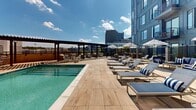 Located at 51 S Broadway in the heart of White Plains, NY, this outdoor pool area offers a resort-inspired retreat set against a vibrant urban skyline. The centerpiece is a generously sized swimming pool with crystal-clear water, flanked by rows of cushioned lounge chairs adorned with classic navy-and-white striped accents, inviting residents to relax and soak in the open sky above.

The deck is finished in light wood-toned porcelain tile that lends a warm, contemporary aesthetic throughout the space. Two fully equipped outdoor kitchen islands serve as focal points for the terrace areas, each featuring stainless steel grills, marble-topped counters, built-in sinks, and bar seating — ideal for al fresco entertaining. Striped patio umbrellas provide shade over multiple dining and lounge table arrangements, creating distinct social zones within the space.

A pergola structure at one end of the pool deck adds architectural character while offering a shaded gathering spot. Sleek dark metal fencing and privacy screens frame the perimeter, balancing openness with a sense of enclosure. The surrounding multi-story building facade, with its modern paneling and private balconies, contributes to the cohesive architectural design. The entire space is bathed in abundant natural sunlight, with sweeping views of the White Plains skyline completing the experience.