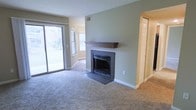This single-floor space offers a comfortable and functional layout with a warm, inviting ambiance. The main living area is finished with plush wall-to-wall carpeting and is anchored by a classic fireplace featuring a dark tile surround and a rich wood mantel, making it a natural focal point of the room. Soft, neutral green walls complement the light-toned trim and baseboards, creating a calm and cohesive atmosphere throughout.

Natural light is a defining feature of this space, courtesy of a full-width sliding glass door that opens to an outdoor view, as well as an additional side window that together bathe the interior in bright, diffused daylight. The sliding door provides convenient outdoor access and enhances the sense of openness within the living area.

A visible hallway with a paneled door leads to additional areas of the home, suggesting a practical and well-organized floor plan. Overhead ventilation is integrated discreetly into the upper portion of the wall, contributing to the clean, uncluttered aesthetic. The combination of the fireplace, generous natural light, and neutral finishes gives this space a timeless, move-in-ready character suited to a variety of interior styles.