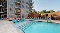 Located at 2300 N Davidson St in Charlotte's NoDa neighborhood, this amenity space offers a thoughtfully designed environment for both relaxation and socializing. The outdoor area showcases a sparkling pool surrounded by a generously sized deck, lined with teal lounge chairs and striped cabana-style seating, all set against the backdrop of a modern multi-story building with balconies and mature trees.

Inside, the single-floor amenity space makes a bold design statement. The main lounge features soaring ceilings with a dramatic dark finish, exposing industrial elements while recessed and pendant lighting add warmth throughout. The signature hexagonal tile flooring in a multi-tone gray, black, and orange palette anchors the space with a striking visual identity. Seating areas are furnished with plush sofas and accent chairs arranged around coffee tables, complemented by colorful, geometric-patterned area rugs and eclectic pendant light clusters.

A dedicated clubroom/kitchen area provides a large dark stone communal table with bar-height seating, a full wet bar with stainless steel appliances, and a wall-mounted TV — ideal for gatherings and events. Large windows and glass garage-style doors allow natural light to flow freely, creating a seamless connection between the interior spaces. Vibrant artwork and textured wall accents complete the energetic, contemporary atmosphere throughout.