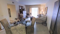 This thoughtfully designed single-floor residence offers a welcoming and cohesive living environment anchored by an open-concept layout. The dining area features a round glass-top table with elegantly upholstered chairs, creating an intimate setting for meals and gatherings. Decorative accents, including layered place settings and tasteful centerpieces, add a refined touch to the space.

Flowing naturally from the dining area, the living room is furnished with a plush, neutral-toned sofa adorned with decorative pillows and a fringed throw, complemented by coordinating accent chairs. A classic armoire and framed wall art contribute to the room's traditional charm. Soft, wall-to-wall carpeting runs throughout, adding warmth and comfort underfoot.

A large sliding glass door floods the living area with natural light, offering an inviting connection to the outdoors while brightening the neutral color palette of beige and warm earth tones. Sheer curtains frame the door, softening the natural light and enhancing the cozy ambiance.

The overall aesthetic is classic and carefully curated, with consistent warm tones, layered textures, and decorative details that create a harmonious and comfortable atmosphere throughout the space.
