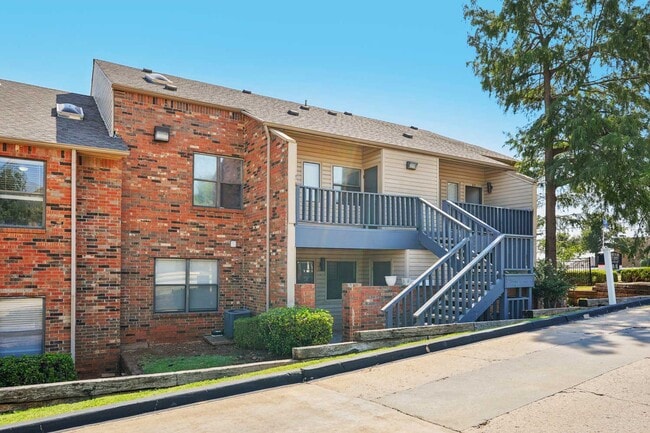 Two-story apartment building exterior with red brick facade, blue staircase, upper balcony, shrubs, and tall trees - Walnut Creek