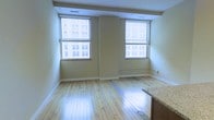 This single-floor space offers a clean, open layout characterized by light-toned hardwood floors that run throughout, reflecting natural light and contributing to an airy, welcoming atmosphere. Two generously sized windows dominate the main living area, framing views of the surrounding urban streetscape and allowing abundant daylight to fill the room. The windows are fitted with horizontal blinds, offering flexible light control. Neutral beige walls complement the warm wood flooring, creating a cohesive and understated aesthetic. Ceiling-mounted HVAC vents suggest a modern climate control system integrated discreetly into the architecture. A partial view of a granite or stone-topped counter at the edge of the frame hints at a kitchen area adjacent to the main living space, featuring cabinetry in a natural wood finish. The overall ambiance is bright and functional, with an open feel well-suited to urban living. The layout appears to flow naturally between the living and kitchen areas, making efficient use of the available space while maintaining a sense of openness throughout.