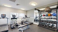Located at 5870 56th Ave N in Kenneth City, Florida, this community amenity space offers residents a well-rounded fitness and recreational experience. The fitness center is housed in a single-story building with a distinctive teal metal roof and a welcoming blue entry door, framed by colorful landscaping. Inside, the gym features durable rubber flooring, flush-mounted ceiling lights, and large windows that allow natural light to fill the space. Equipment includes treadmills, an elliptical, a stationary bike, a cable machine, a weight bench, and a well-stocked dumbbell rack alongside medicine balls, providing a comprehensive workout environment. A full-length mirror expands the sense of space and aids in exercise form. Adjacent to the fitness center, the outdoor area showcases a sparkling pool with decorative tile accents, surrounded by a paver brick deck ideal for lounging. Wrought iron fencing encloses the pool area, and poolside seating offers a relaxing retreat. Towering royal palms and mature tropical trees create a lush, resort-like atmosphere throughout the grounds. Meandering concrete walkways wind through manicured lawns and vibrant garden beds, connecting the multi-story residential buildings and enhancing the overall appeal of this well-maintained Florida community.