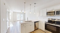 Located at 201 Munn Rd in Nashville, TN, this single-floor unit offers a thoughtfully designed layout that maximizes functionality within a compact footprint. The open-concept main area seamlessly integrates the kitchen, dining, and living spaces, creating a bright and cohesive environment. The kitchen is well-appointed with white shaker-style cabinetry, a subway tile backsplash, a peninsula with an undermount sink and pendant lighting overhead, and a full suite of stainless steel appliances including a refrigerator, range, microwave, and dishwasher. Light wood-style flooring runs throughout the main living areas, complementing the neutral, warm color palette. The living area benefits from abundant natural light streaming through large windows and a set of French doors that open to a private balcony, offering views of the surrounding greenery and neighborhood streetscape. Ceiling fans are present in both the living area and bedroom, enhancing comfort year-round. The bedroom features carpeted flooring and access to two closets, including a walk-in. The bathroom includes a full soaking tub and shower combination with clean, modern finishes. A dedicated laundry area and utility room add everyday practicality to this well-rounded Nashville residence.