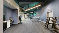 Located at 947 N Park Ave in Tucson, Arizona, this contemporary fitness center offers a well-appointed, single-floor layout designed to support a full range of training activities. The space features an open, industrial-style aesthetic with exposed ductwork, teal-painted ceilings, and concrete structural columns that lend a modern, urban character throughout.

The main workout floor is divided into distinct functional zones. The cardio area lines one wall with a row of treadmills, stair climbers, and stationary bikes, all positioned beneath warm linear pendant lighting and a vibrant neon "Feeling Sharp" sign that adds a lively, motivating energy to the space. Adjacent to this, a strength training zone features a selection of cable machines, selectorized equipment, and benches, all set on bright green turf flooring that clearly delineates the area. A free weights section offers a comprehensive rack of dumbbells, kettlebells, medicine balls, and sandbags.

Large mirrors are strategically placed throughout to enhance the sense of space and support proper form. A separate, enclosed studio room features light wood flooring, a wall-mounted flat-screen television, and a mounted display, making it well-suited for group classes or personal training sessions. The flooring throughout the main area transitions between patterned gray carpet and turf, providing both comfort and performance underfoot. The overall ambiance is energetic yet polished, reflecting a professionally designed fitness environment.