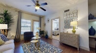 This single-story unit offers a thoughtfully designed layout that connects the main living spaces seamlessly. The living area features warm hardwood-style flooring, a ceiling fan, and natural light from windows dressed with blinds and drape panels. A bold patterned area rug anchors the seating arrangement, complemented by eclectic wall décor and a mirrored dresser that adds a touch of elegance.

The kitchen is outfitted with dark espresso cabinetry, granite countertops, and stainless steel appliances including a refrigerator and microwave. A curved kitchen island with a built-in wine rack doubles as a breakfast bar with bar stool seating, making it both functional and stylish. An adjacent built-in desk or display shelf adds additional utility to the space.

The bedroom is a serene retreat with plush carpeting, a ceiling fan, a natural-light window flanked by framed artwork, and ample space for generous furnishings.

The bathroom features dark wood cabinetry with a wide granite-topped vanity, a large mirror, and a full soaking tub with a tile surround. Hardwood-style flooring continues into this space, reinforcing the cohesive design throughout the unit.
