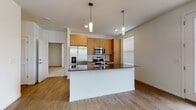 This single-floor unit in Kenosha, Wisconsin, showcases a clean, modern design with an open-concept layout that maximizes the sense of space and flow. The kitchen serves as a central highlight, featuring warm honey-toned wood cabinetry, a tile backsplash, and a full suite of stainless steel appliances, including a French door refrigerator, range, and built-in microwave. A substantial center island with a dark countertop and pendant lighting above provides both prep space and a natural gathering point. The kitchen opens seamlessly into a generously sized living area finished with light wood-tone flooring, creating a bright and cohesive atmosphere. A glass exterior door allows natural light to fill the space while offering views of the surrounding neighborhood. The bedroom features plush neutral carpeting and connects to a walk-in closet outfitted with wire shelving and hanging rods for efficient storage. The bathroom is finished consistently with the rest of the unit, offering a wood-toned vanity, dark countertop, large mirror, and matching flooring. An attached garage with an automatic opener provides convenient, secure parking and additional storage space, completing this well-appointed, move-in-ready unit.