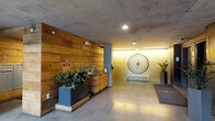 This single-floor space presents a thoughtfully designed interior that blends industrial and contemporary aesthetics. The lobby area immediately draws attention with its exposed concrete ceiling and polished concrete floors, creating a raw yet refined atmosphere. A prominent feature wall clad in warm, horizontal wood planks anchors one side of the space, adding natural texture and warmth that contrasts beautifully with the cooler concrete surfaces.

At the far end of the lobby, a vivid yellow brick wall serves as a dramatic focal point, illuminated by ambient lighting that enhances its bold color. Mounted centrally on this wall is a striking circular lightning bolt emblem, lending the space a distinctive branded identity. Recessed and pendant lighting throughout ensures the area remains well-lit while contributing to the overall ambiance.

A reception or service counter, finished in matching wood tones, is positioned along the wood-paneled wall and is accompanied by lush potted plants in modern charcoal planters, softening the industrial elements with organic greenery. A floor mat near the entrance adds a practical finishing touch. Overall, the space communicates a confident, urban character with carefully balanced materials and design details.