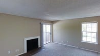 This single-floor space centers around a comfortable living area finished with wall-to-wall carpet and neutral-toned walls that create a warm, understated atmosphere. A fireplace with a clean white surround and dark insert serves as a focal point along one wall, adding both character and function to the room. The textured popcorn ceiling runs throughout, lending a classic residential feel to the interior. A sliding glass door floods the space with natural light and opens directly onto a private balcony, providing an outdoor extension of the living area with views of the surrounding exterior. The transition between the carpeted living space and the tiled area near the fireplace adds subtle visual definition to the room. The overall layout is open and straightforward, offering a clean canvas that is easy to furnish and personalize. White baseboards frame the perimeter of the room, adding a crisp contrast to the neutral walls and flooring. The space presents a practical and functional design, well-suited for everyday living.