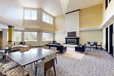 Located at 6000 NW 62nd Ave in Johnston, Iowa, this expansive single-floor community space offers a sophisticated and welcoming environment designed for gathering and relaxation. The centerpiece of the main lounge is a dramatic floor-to-ceiling textured stone fireplace flanked by a mounted flat-screen TV, surrounded by sleek dark leather seating arranged on a neutral area rug. Soaring double-height ceilings amplify the sense of grandeur, and a cascading crystal chandelier adds an elegant focal point overhead. Abundant natural light pours in through large windows lining multiple walls, offering views of the outdoor pool and surrounding landscaping. The lounge transitions seamlessly into multiple dining and casual seating areas, including high-top bistro tables positioned near the windows for intimate gatherings. The fully equipped kitchen features rich dark cabinetry, granite countertops, light hardwood flooring, stainless steel appliances — including a refrigerator, double wall oven, and dishwasher — and a spacious central island with bar seating illuminated by pendant lighting. Decorative upper windows add architectural interest throughout the space. An ADA-compliant bathroom with grab bars and tile flooring provides accessible facilities. The overall ambiance is modern, polished, and community-oriented.