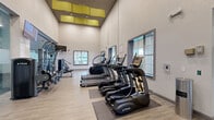 Located at 2801 Biltmore Park Dr in Orlando, FL, this well-appointed fitness center spans a generous single floor, offering a comprehensive range of workout environments designed to support diverse fitness goals. The facility is divided into three distinct zones, each thoughtfully designed to serve a specific training purpose.

The main cardio and machine area features soaring ceilings with eye-catching yellow acoustic panel accents, light wood-style flooring, and an array of premium TRUE-brand treadmills, ellipticals, and cable machines. Natural light filters in through large windows, enhancing the open, energizing atmosphere. Wall-mounted flat-screen TVs provide entertainment throughout.

A dedicated functional training zone, separated by sleek floor-to-ceiling glass partition walls, offers rubber flooring, power racks with free weights, battle ropes, air bikes, rowing machines, and large wall-mounted mirrors — all complemented by dramatic fitness-themed mural artwork. Ceiling fans and additional TVs complete this high-performance space.

A separate yoga and flexibility studio provides a calm contrast, featuring plush carpet flooring, elegant globe pendant lighting, and a serene full-wall mural of a meditating figure at sunset, creating an ideal environment for yoga, stretching, and mindfulness practices. Together, the three areas form a cohesive, full-service fitness facility.