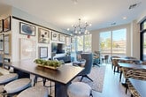 Located at 375 Gartrell St SE in Atlanta's vibrant Grant Park neighborhood, this thoughtfully designed amenity space combines comfort, functionality, and contemporary style across a single floor. The combined living and dining area serves as the heart of the space, where light wood-look flooring, large floor-to-ceiling windows, and French doors flood the interior with abundant natural light and offer open views of the surrounding landscape. A striking sculptural chandelier anchors the dining zone, which features a substantial dark-topped communal table with bar-height seating. The living area is anchored by a gallery wall of curated artwork surrounding a wall-mounted flat-screen TV, complemented by plush seating in tones of teal, gray, and blue arranged over a patterned area rug. Cafe-style tables near the windows provide additional flexible seating. A wet bar with navy cabinetry and white countertops adds a refined, functional touch. The dedicated office space offers a vibrant contrast, highlighted by a bold multicolored vertical slat accent wall in red, yellow, green, and blue. This room is equipped with a conference table, ergonomic task chairs, a wall-mounted display screen, a whiteboard, and a kitchenette with gray cabinetry and subway tile backsplash — making it an ideal setting for focused work or collaborative meetings.