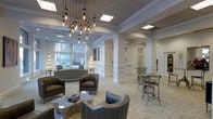 This refined single-floor space combines classic architectural details with contemporary furnishings to create an inviting and polished atmosphere. The interior is defined by crisp white walls adorned with traditional wainscoting panels and punctuated by decorative columns that add a sense of structure and depth throughout the open layout.

Natural light streams in generously through large street-facing windows at the front of the space, offering views of the surrounding streetscape and creating a bright, airy feel. Overhead, a dramatic cluster chandelier featuring exposed Edison-style bulbs serves as a striking focal point, complementing the recessed and flush ceiling lighting distributed across the paneled ceiling.

The seating areas are thoughtfully arranged and furnished with gray leather armchairs and a matching sofa accented with nailhead trim, centered around an organic-shaped coffee table. Toward the rear, bistro-style high-top tables paired with cross-back bar stools offer a more casual seating option. Display fixtures and decorative accents, including tiered stands likely showcasing merchandise, are visible along the perimeter, suggesting a retail or hospitality-oriented function. The overall ambiance is elegant, light-filled, and professionally curated.