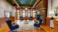 Located at 1000 Flycatcher Way in Arden, NC, this single-story craftsman-style space exudes warmth and sophistication through its thoughtfully curated architectural details. Rich hardwood floors stretch throughout the open layout, complemented by extensive warm-toned wood trim, wainscoting, and millwork that frame every doorway and window. The centerpiece of the main gathering area is a dramatic floor-to-ceiling stacked stone fireplace, complete with a rustic wood mantel, decorative ironwork screen, and a raised stone hearth — creating an inviting focal point for the entire room. Above, a stunning coffered ceiling painted in deep teal adds a bold contrast against the natural wood beams and white crown molding, while a wrought iron chandelier adds an elegant finishing touch. Expansive windows with transoms on multiple walls flood the space with natural light and offer views of the surrounding greenery, reinforcing the connection to the North Carolina landscape. A comfortable seating arrangement with wingback chairs and a central coffee table makes this an ideal gathering space. Additional seating nooks near the windows provide quieter retreats. The overall ambiance blends rustic mountain charm with refined craftsmanship, making this a welcoming and visually impressive community space.