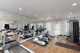 This fitness center is designed to support a well-rounded workout routine in a bright, welcoming environment. The space features warm wood-look flooring throughout, neutral gray walls with clean white trim, and recessed lighting complemented by natural light streaming through multiple white-shuttered windows. A dedicated exterior door provides convenient direct access.

A large floor-to-ceiling mirror wall visually expands the space and allows users to monitor their form during exercise. The cardio section is comprehensively outfitted with multiple TRUE-brand treadmills and elliptical cross-trainers, positioned to take advantage of natural light and mirror sightlines. An upright stationary bike rounds out the aerobic options.

For strength training, the facility includes an Inspire multi-functional cable machine, a weight bench, and a cable/pulley system, offering a solid variety of resistance training options. A stability ball and kettlebell are also visible, supporting functional fitness and flexibility work. Exercise mats are available on the floor for stretching and bodyweight exercises.

Safety features include a fire extinguisher, an emergency exit sign, and posted fitness center rules. The space is thoughtfully laid out to allow comfortable movement between equipment stations, making it functional for multiple users at once.