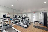 This fitness center is designed to support a well-rounded workout routine in a bright, welcoming environment. The space features warm wood-look flooring throughout, neutral gray walls with clean white trim, and recessed lighting complemented by natural light streaming through multiple white-shuttered windows. A dedicated exterior door provides convenient direct access.

A large floor-to-ceiling mirror wall visually expands the space and allows users to monitor their form during exercise. The cardio section is comprehensively outfitted with multiple TRUE-brand treadmills and elliptical cross-trainers, positioned to take advantage of natural light and mirror sightlines. An upright stationary bike rounds out the aerobic options.

For strength training, the facility includes an Inspire multi-functional cable machine, a weight bench, and a cable/pulley system, offering a solid variety of resistance training options. A stability ball and kettlebell are also visible, supporting functional fitness and flexibility work. Exercise mats are available on the floor for stretching and bodyweight exercises.

Safety features include a fire extinguisher, an emergency exit sign, and posted fitness center rules. The space is thoughtfully laid out to allow comfortable movement between equipment stations, making it functional for multiple users at once.