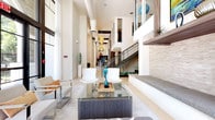 This expansive, single-floor communal space showcases a striking contemporary design defined by soaring double-height ceilings and an open, flowing layout that connects multiple distinct zones seamlessly. The main lounge area draws immediate attention with its dramatic vertical scale, featuring sculptural pendant lighting, crisp white walls, and large floor-to-ceiling windows dressed in sheer curtains that flood the interior with natural light and offer views of the surrounding greenery outside.

Throughout the space, carefully curated artwork and decorative sculptures add personality and visual interest, complementing the refined neutral palette of light tile flooring and textured stone accent walls. Seating areas are thoughtfully arranged, offering a mix of sleek modern chairs, plush leather sofas with warm accent cushions, and glass-topped coffee tables — creating inviting settings for both casual conversation and focused gatherings.

A dedicated lounge section features a wall-mounted flat-screen television, stone-clad accent walls with warm underlighting, and rich brown leather seating arranged around a central coffee table. The kitchenette is well-appointed with warm wood cabinetry, light stone countertops, a blue glass tile backsplash, under-counter refrigeration, and a large communal dining island illuminated by colorful pendant lights. A package hub station adds a practical modern convenience. A staircase with sleek metal railings connects to an upper mezzanine level, adding architectural depth to this beautifully designed space.