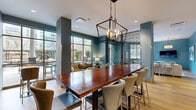 This well-appointed amenity lounge is a thoughtfully designed communal space that seamlessly blends multiple functional zones within a single, open floor plan. The interior is unified by a warm hardwood floor and a consistent teal color palette across textured walls, creating a cohesive and inviting atmosphere throughout.

A central highlight is the long communal dining table crafted from rich, dark wood, surrounded by upholstered bar-height chairs and anchored above by a striking geometric lantern-style chandelier. Adjacent to this gathering area, a smaller round table with elegantly detailed teal chairs sits beside a glass door that opens directly to an outdoor terrace, filling the corner with natural light and greenery views.

A dedicated lounge area features a curated arrangement of plush sofas, swivel accent chairs in soft aqua tones, and a low-profile coffee table, all oriented around a wall-mounted television and complemented by framed artwork. Floor-to-ceiling windows along one wall flood this seating zone with abundant natural light while offering panoramic views of an outdoor courtyard and the surrounding cityscape.

Decorative lattice screens serve as elegant architectural dividers between zones, adding visual depth without sacrificing openness. High bistro tables with gold-accented chairs provide additional flexible seating. The overall ambiance is polished yet comfortable, balancing contemporary design elements with warm, welcoming furnishings ideal for both socializing and relaxing.