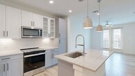 This well-appointed 1-bedroom, 1-bathroom unit offers a thoughtfully designed layout that maximizes both comfort and functionality. The open-concept kitchen and living area serves as the heart of the home, showcasing a large center island with a sleek quartz countertop, an undermount sink, and stylish pendant lighting overhead. The kitchen is equipped with stainless steel appliances — including a French door refrigerator, electric range, built-in microwave, and dishwasher — complemented by two-tone cabinetry in white and soft grey with glass-front upper cabinets for a refined touch. White subway tile backsplash and recessed lighting add to the clean, contemporary aesthetic. Light wood-style flooring runs throughout the main living areas and bedroom, lending warmth and continuity. The living area features a ceiling fan and glass French doors that open to a private balcony, bringing in abundant natural light. The spacious bedroom offers a ceiling fan and easy access to the full bathroom, which features a dual-sink vanity with a dark countertop, tile flooring, a walk-in shower, and direct access to a generously sized walk-in closet with built-in shelving and hanging rods.