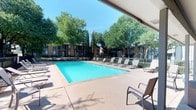 This outdoor amenity space centers around a generously sized rectangular swimming pool with clear, vivid turquoise water, designed to serve as a premier recreational focal point for the surrounding residential community. The pool deck is finished in a neutral concrete surface and offers ample room for lounging, lined on multiple sides with rows of sling-style lounge chairs and seating arrangements ideal for relaxation and sunbathing. A covered patio area along one side of the deck provides shaded respite from the sun, extending the usability of the space throughout the day. The pool area is enclosed by a classic black iron fence, offering both safety and a clean, defined boundary. Towering mature trees — including large shade trees and slender cypress varieties — surround the perimeter, providing a lush, green backdrop and natural cooling shade. The adjacent clubhouse or amenity building features a pitched roof with warm brick exterior, complementing the traditional architectural style of the multi-story residential buildings visible in the background. The overall ambiance is open, bright, and inviting, with abundant natural light and a tranquil, park-like atmosphere well-suited for both active recreation and leisurely outdoor enjoyment.