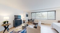 This single-floor studio is thoughtfully designed to maximize both function and comfort through an efficient open-plan layout. The main living area serves as the heart of the space, featuring light hardwood flooring throughout and crisp white walls that create a clean, airy atmosphere. A large picture window dominates one wall, flooding the room with natural light and offering a lush, tree-lined view that brings the outdoors in. The living zone accommodates a comfortable sofa, a glass-top coffee table, and a media console, while a compact dining area with a round table and chairs sits conveniently beside the window.

The sleeping area flows naturally from the living space, providing room for a full-size bed and a built-in closet with open shelving, offering practical storage solutions. The kitchen is efficiently appointed with stainless steel appliances, including a gas range, over-the-range microwave, and refrigerator, complemented by white cabinetry and dark stone countertops with a tile backsplash. The full bathroom maintains the space's clean aesthetic, featuring a white vanity with dark stone countertop, a tub-shower combination with bright white tile surround, and light tile flooring. Overall, the space presents a modern, well-organized environment with consistent finishes throughout.