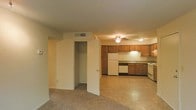 This single-floor unit features an open-concept layout that seamlessly connects the living area to the kitchen, creating a sense of spaciousness throughout. The living area is finished with wall-to-wall carpeting, providing a warm and comfortable underfoot feel, while a ceiling fan overhead aids in air circulation. A built-in closet with a hanging rod is conveniently positioned near the living space, offering practical storage solutions.

The kitchen transitions from the carpeted area via light-toned vinyl flooring and is equipped with warm wood-toned cabinetry that extends along the walls, providing ample storage. White appliances, including a refrigerator, dishwasher, and stovetop, are neatly integrated into the cabinetry. An under-cabinet range hood sits above the cooking area, and a sink is positioned along the counter with additional workspace on either side. The kitchen's ceiling fan provides additional ventilation. Neutral-toned walls throughout the unit contribute to a clean, understated aesthetic, while HVAC vents in the ceiling ensure consistent climate control throughout the space.