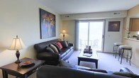 This single-floor space offers a welcoming and thoughtfully designed living area that balances comfort with style. The living room serves as the centerpiece of the space, featuring plush leather sofas arranged around a sleek coffee table, creating an inviting setting for relaxation and entertaining. Soft, neutral-toned carpeting adds warmth underfoot, complementing the light-colored walls that lend the room an airy, open feel. A large sliding glass door floods the room with abundant natural light and opens directly onto a private balcony, where outdoor seating allows for a seamless indoor-outdoor living experience. The balcony view adds a sense of openness and connectivity to the outdoors. A decorative floor lamp provides warm ambient lighting, while a bold, colorful floral artwork displayed prominently on the wall adds a vibrant focal point and a touch of artistic character to the room. Subtle architectural details, including a ceiling vent and clean wall lines, reflect a practical yet polished design. Overall, the space presents a comfortable, light-filled environment well-suited for everyday living.