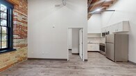 Located at 724 N Harrison St in Ludington, Michigan, this single-floor space masterfully combines industrial heritage with modern design. The main living area commands attention with dramatically soaring ceilings, exposed wooden ceiling beams, and a striking exposed brick accent wall that adds texture and warmth. Light-toned luxury vinyl plank flooring runs throughout, complementing the crisp white walls and dark baseboards for a clean, contemporary aesthetic. A bold black structural column anchors the open layout, serving as a nod to the building's architectural past.

The kitchen is well-appointed with white shaker-style cabinetry extending to the ceiling for ample storage, granite countertops, and a classic white subway tile backsplash. Stainless steel appliances — including a refrigerator, electric range, built-in microwave, and dishwasher — complete the space with a polished, functional finish.

The bathroom continues the modern aesthetic with a white shaker vanity topped with granite, a framed mirror, stylish pendant lighting, and wainscoting detail along the lower walls. An additional room accessible through a doorway from the main living area provides flexible use of space. Industrial-style ductwork adds to the character of this thoughtfully renovated property.