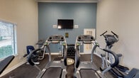 Located at 1920 W Tarrant Rd in Grand Prairie, Texas, this fitness center is a single-floor facility designed to support a well-rounded workout routine. The space features a two-tone color scheme with a calming slate-blue accent wall complemented by neutral beige and off-white walls, giving it a clean and focused atmosphere. Crown molding and white baseboards add a polished architectural touch throughout.

The room is equipped with multiple treadmills, an elliptical trainer, and a stair climber, providing a comprehensive range of cardio options. A three-tier dumbbell rack holds a full set of free weights, accommodating strength training needs. A weight bench completes the free weight area. A wall-mounted flat-screen television is centrally positioned for entertainment during workouts, and a paper towel dispenser and sanitation station promote cleanliness.

Natural light filters in through slatted window blinds along one wall, offering views of the surrounding outdoor greenery while maintaining privacy. Full-length mirrors line one wall, visually expanding the space and allowing users to monitor their form. Dark rubber flooring throughout provides durability and traction. The room is accessed through a French-style glass-panel door, and ceiling-mounted flush lighting ensures the space remains well-illuminated at all times.