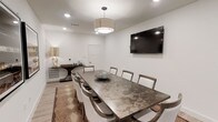 This single-floor space centers around a well-appointed conference or dining room designed for both functionality and style. The room features a large, rectangular table with a distinctive metallic, mottled finish that serves as the focal point, surrounded by upholstered chairs in neutral tones that offer both comfort and sophistication. A decorative bowl centerpiece adds a subtle artistic touch to the table surface.

Overhead, a modern drum pendant light provides warm, diffused illumination, complemented by recessed ceiling lighting that ensures the room is evenly lit. A large wall-mounted flat-screen television is positioned prominently on one wall, making the space well-suited for presentations or media viewing.

The walls are finished in a clean, neutral white, contributing to a bright and open atmosphere. Light wood-tone flooring runs throughout, grounded by a patterned area rug beneath the table that adds visual warmth and texture. Along one wall, a dark wood console or credenza provides additional surface and storage space, accented by a sculptural lamp.

The overall ambiance is polished and contemporary, balancing professional utility with refined interior design details.