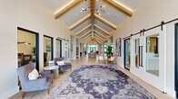 Located at 2300 SW 38th Ave in Ocala, Florida, this generously sized single-story community clubhouse offers an impressive and thoughtfully designed gathering space. The architectural centerpiece is a dramatic vaulted ceiling adorned with warm wood beams and integrated ambient lighting, creating a grand yet welcoming atmosphere throughout the main corridor and living areas.

The space features two distinct living rooms, each with its own character. One showcases floor-to-ceiling black-framed windows that flood the room with natural light and offer views of the outdoor amenity area, complemented by elegant drapery and curated seating arrangements. The other living area is anchored by a white brick fireplace with a mounted TV, teal built-in shelving, and mid-century modern furnishings arranged over patterned area rugs.

Light hardwood flooring runs seamlessly throughout, enhancing the bright, airy feel. A dedicated lounge and dining nook with booth-style seating and patterned tile accents provides a more intimate gathering spot. Sliding barn doors, bold artwork, and lush indoor greenery add refined touches. Large glass doors connect the interior to an outdoor patio, blending indoor and outdoor living effortlessly.
