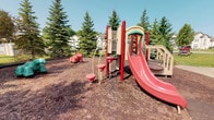 Located at 842 Cobblestone Cir in Painesville, Ohio, this outdoor recreational space offers a welcoming playground area designed for active play. The centerpiece of the space is a multi-component play structure finished in red and tan, featuring a curved slide, climbing elements, and a spring-mounted animal ride, all set atop a generous wood chip safety surface that cushions the play area. Tall, mature evergreen trees frame the perimeter of the playground, providing a lush green backdrop and natural shade during warmer months. The surrounding landscape creates a sense of enclosure and privacy while maintaining an open, airy feel under the clear blue sky. Residential buildings are visible through the treeline, indicating the playground's role as a shared community amenity. The space is well-suited for young children, offering a variety of interactive features in a safe and inviting outdoor setting. The combination of vibrant play equipment, natural landscaping, and a thoughtfully maintained ground surface makes this playground a functional and appealing gathering spot within the community.