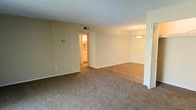 This single-floor residence offers a thoughtfully arranged layout that emphasizes open, connected living spaces. The main living area features plush wall-to-wall carpeting underfoot and is finished with neutral beige walls and a textured ceiling, creating a warm and understated ambiance. A thermostat on the wall speaks to the practical functionality built into the space.

Transitioning naturally from the living area, the dining space is distinguished by a change in flooring to smooth hardwood-style planks, visually defining the two zones while maintaining an open, flowing feel. A pendant light fixture hangs above, adding a refined focal point to the dining area.

A doorway leads directly into the kitchen, which is visible in the background and features light cabinetry, offering a practical and accessible cooking space that connects seamlessly to the rest of the home. The HVAC vent positioned on the wall ensures consistent climate control throughout. Bi-fold closet doors are visible near the dining area, providing convenient built-in storage. Overall, the space presents a clean, functional, and comfortable environment well-suited for everyday living.