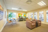 Located at 832 Blackfriars Dr in Virginia Beach, Virginia, this single-floor community amenity space is designed to offer residents a comfortable and functional environment for both work and relaxation. The space is thoughtfully organized into distinct functional zones that flow seamlessly into one another.

The business center area features dedicated workstations equipped with computers and printers, set against warm neutral walls adorned with vibrant, colorful circular artwork that adds an energetic focal point. A glass door provides a view to the outdoors, allowing natural light to brighten the workspace. Large indoor plants contribute a fresh, inviting atmosphere to the area.

Adjacent to the business center is a well-appointed lounge and gathering area, furnished with round tables and cushioned chairs in warm mustard and earth tones. A wall-mounted flat-screen television and decorative sculptural wall art enhance the relaxed ambiance, while a brochure rack provides informational resources for residents. Abstract canvas paintings adorn the walls, adding an artistic and sophisticated touch throughout.

A wall of French doors connects the lounge directly to a fitness center, visible through the glass panes, ensuring convenient access to exercise equipment. The space is finished with commercial-grade carpeting, a drop tile ceiling with recessed lighting, and clean white trim throughout, creating a polished and cohesive community environment.
