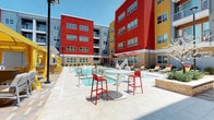 This dynamic outdoor amenity space is defined by bold architectural design, with striking multi-story buildings finished in vivid red, yellow, and gray facades forming an enclosed, resort-style courtyard. At the heart of the space sits a generously sized swimming pool with built-in water fountains, complemented by a separate spa or wading area. The expansive pool deck is finished in light-toned pavers and furnished with chaise lounge chairs, red umbrellas, and high-top bistro tables, creating an inviting atmosphere for relaxation and socializing.

A standout feature is the private poolside cabana, outfitted with comfortable lounge seating, a ceiling fan, a wet bar or beverage station, and drapable curtains for shade and privacy. Adjacent to the pool, a built-in outdoor kitchen features a stainless steel grill set into a natural stone surround, ideal for outdoor entertaining. A dedicated lounge zone offers plush gray sofas accented with rust-colored pillows, centered around an octagonal stone fire pit with a marble top, providing a cozy gathering spot.

The courtyard's textured stone accent walls, patterned paver pathways, tropical landscaping, and cohesive color palette create a lively, resort-inspired ambiance throughout the space.