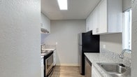 This single-story unit offers a thoughtfully arranged floor plan across two bedrooms and one bathroom. The kitchen is a functional galley-style space featuring white cabinetry with contemporary hardware, granite-look countertops, and stainless steel appliances including an electric range and refrigerator. A skylight overhead floods the kitchen with natural light, enhancing the clean, bright aesthetic. Light-toned hardwood-style flooring runs throughout, adding a cohesive and modern feel underfoot. Textured walls and a popcorn ceiling lend a classic character to the interiors. A hallway connects the main living areas, leading toward a room that benefits from natural light through vertical blinds-covered windows, offering a view of the outdoors. The overall ambiance is clean and move-in ready, with updated finishes that balance practicality and comfort. The layout makes efficient use of its footprint, providing dedicated spaces for cooking, living, and sleeping within a compact but well-organized design.